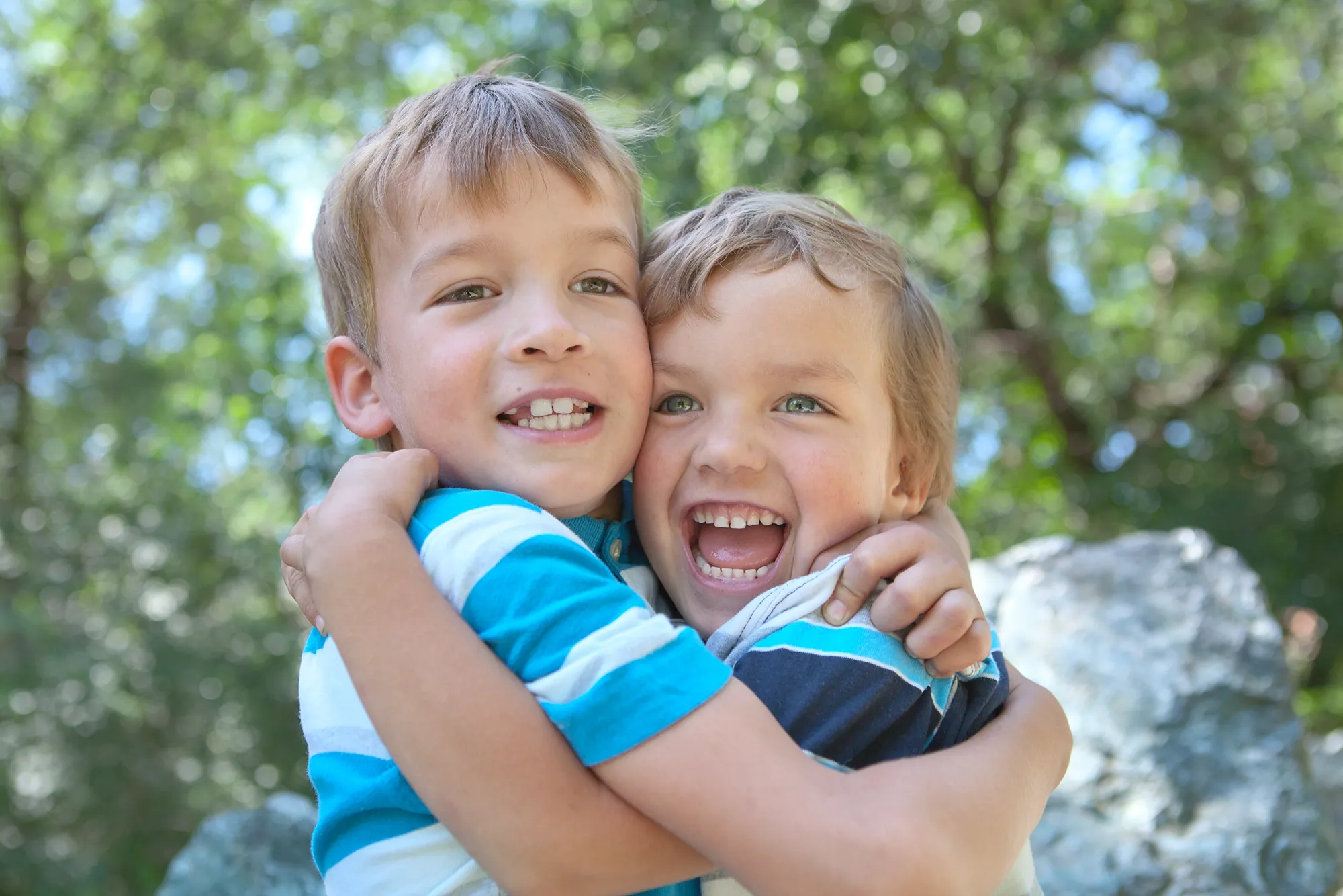 Zwei Jungen umarmen sich lachend im Sommer, im Hintergrund grüne Bäume.