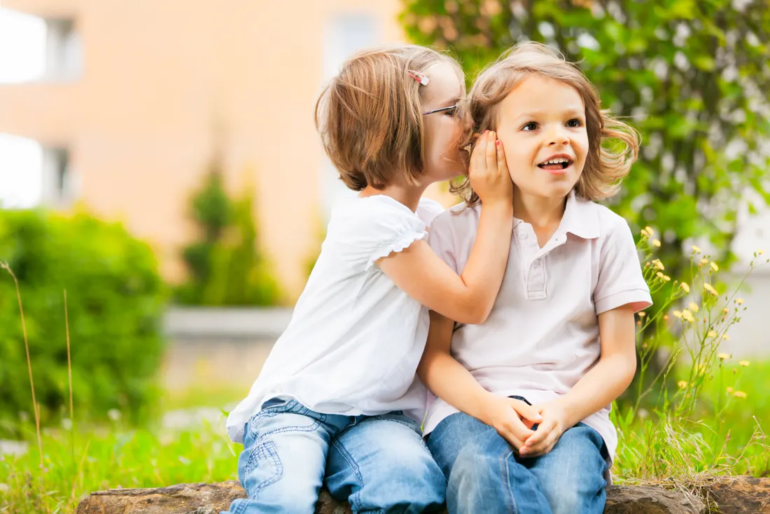 Zwei Kindergartenkinder sitzen im Garten auf einem Stein, eines flüstert dem anderen etwas ins Ohr.
